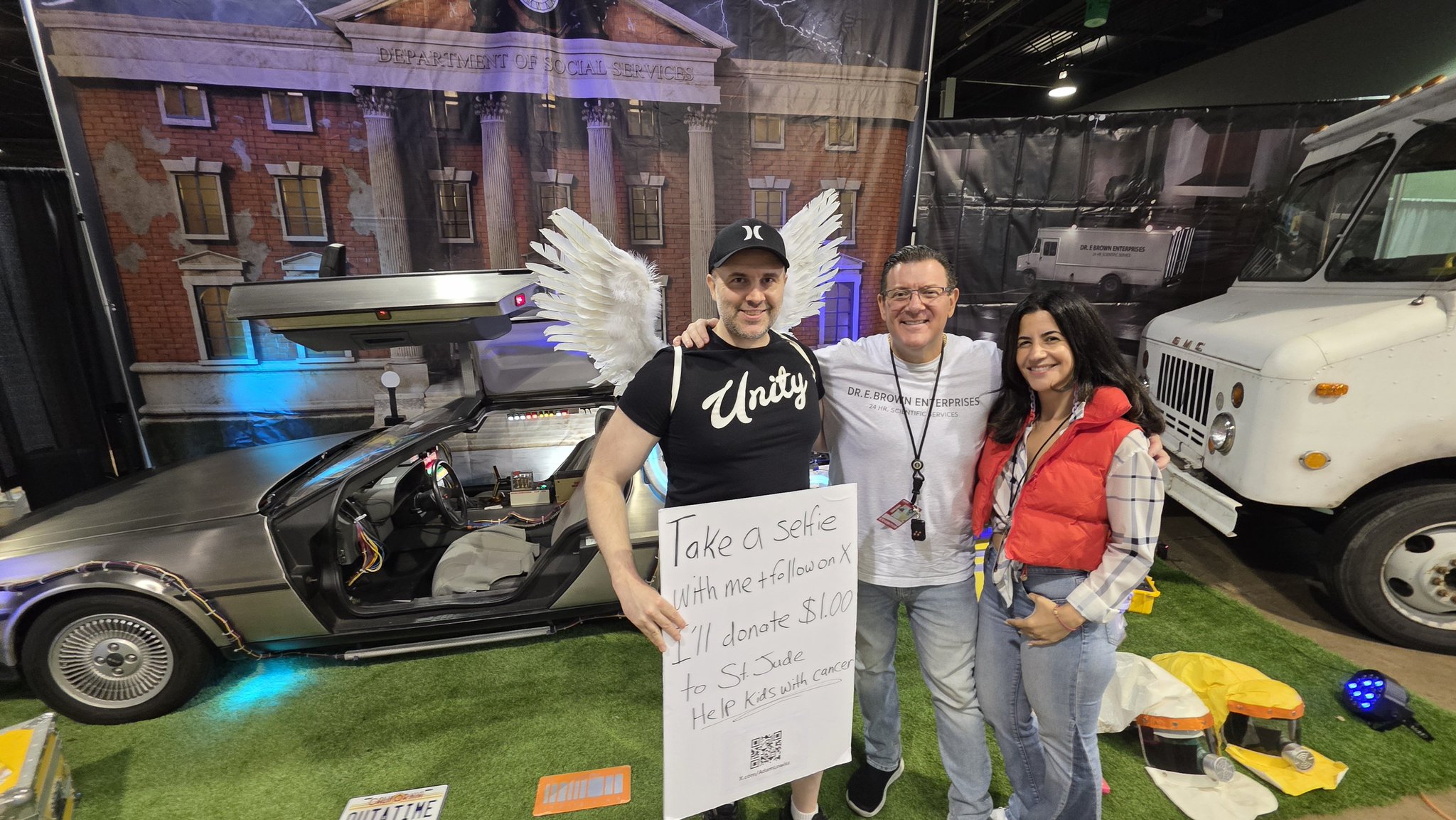 Adam at Fan Expo holding a sign that reads Take a Selfie with me + follow I’ll donate $1, standing beside a DeLorean